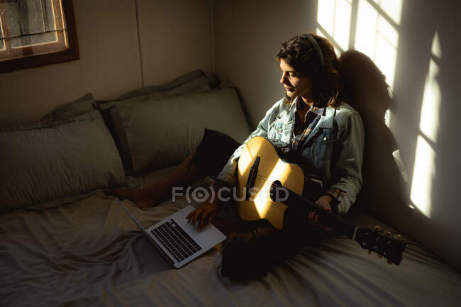 Mulher de raça mista tocando guitarra e usando laptop no quarto ensolarado. estilo de vida saudável, desfrutando de tempo de lazer em casa. — Fotografia de Stock