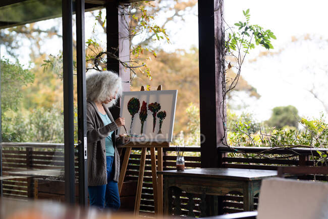 Eine ältere kaukasische Frau steht auf einem sonnigen Balkon und malt. Lebensstil im Ruhestand, Zeit allein zu Hause verbringen. — Stockfoto