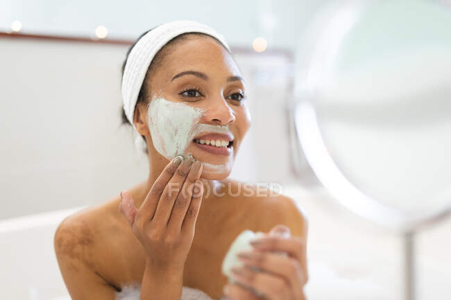 Sorridente donna razza mista in bagno, avendo un bagno e l'applicazione di maschera viso di bellezza. stile di vita domestico, godendo di auto cura del tempo libero a casa. — Foto stock