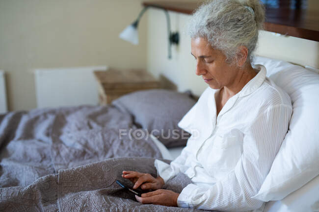 Ältere kaukasische Frau im Schlafzimmer, im Bett sitzend und mit Smartphone. Lebensstil im Ruhestand, Zeit allein zu Hause verbringen. — Stockfoto