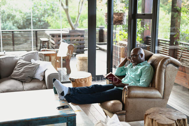 Détendez-vous homme afro-américain senior assis et le livre de lecture dans le salon moderne. mode de vie à la retraite, passer du temps seul à la maison. — Photo de stock