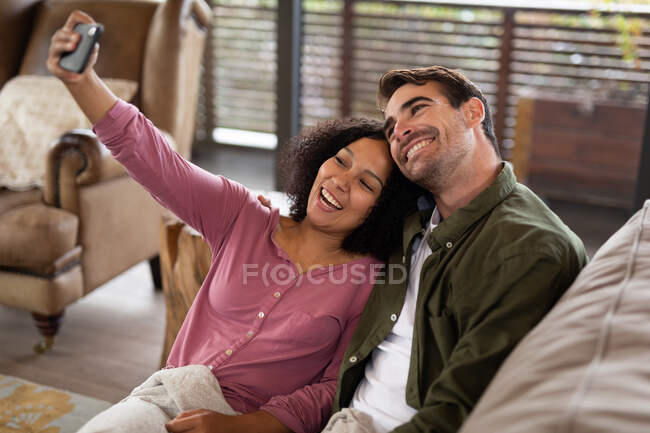 Feliz pareja diversa sentada en el sofá en la sala de estar tomando selfie y sonriendo. pasar tiempo libre en casa en apartamento moderno. - foto de stock