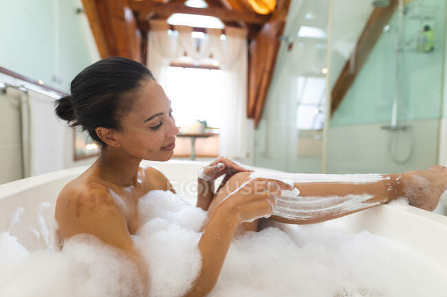 Змішана расова жінка у ванній має ванну і голиться ногами. домашній спосіб життя, насолоджуючись доглядом за дозвіллям вдома . — стокове фото