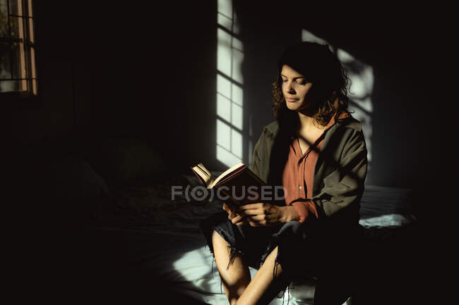 Donna razza mista seduta e lettura libro in camera da letto soleggiata. stile di vita sano, godendo del tempo libero a casa. — Foto stock