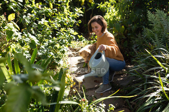 Mujer caucásica sonriente en el jardín con su perro mascota, jardinería. estilo de vida doméstico, disfrutando del tiempo libre en casa. - foto de stock