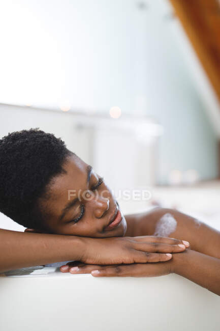 Улыбающаяся африканская американка в ванной, расслабляющаяся в ванне с закрытыми глазами. домашний образ жизни, наслаждаясь отдыхом на дому. — стоковое фото