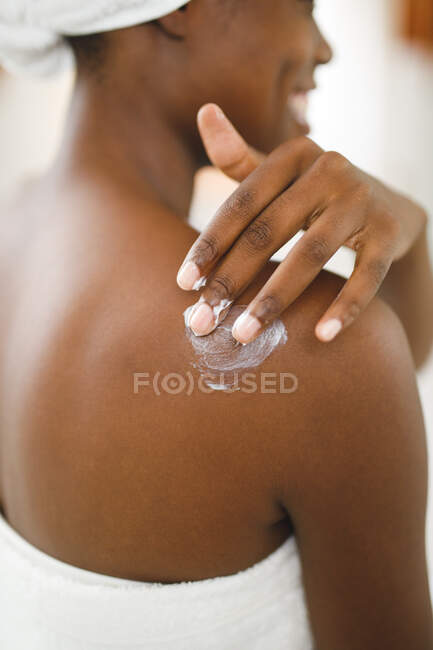 Femme afro-américaine souriante dans la salle de bain appliquant de la crème pour le corps à son épaule pour les soins de la peau. mode de vie domestique, profiter de loisirs d'auto-soins à la maison. — Photo de stock