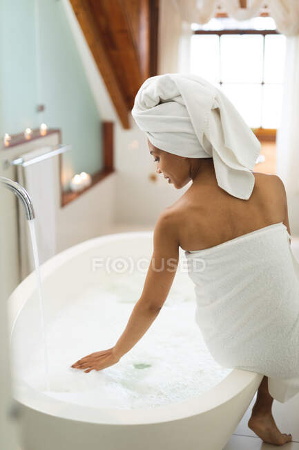 Mixed race woman in bathroom running a bath, sitting on the edge of tub. domestic lifestyle, enjoying self care leisure time at home. — Stock Photo