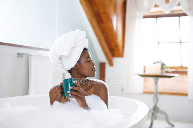 Усміхнена афро-американська жінка у ванній кімнаті, відпочиває у ванній і п'є каву. Домашній спосіб життя, дозвілля для себе вдома. — стокове фото
