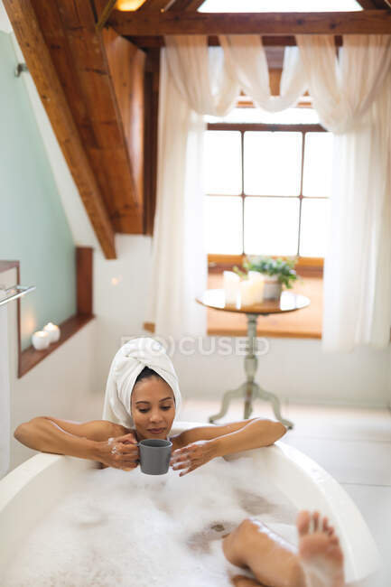 Happy mixed race woman in bathroom having a bath and drinking coffee. domestic lifestyle, enjoying self care leisure time at home. — Stock Photo
