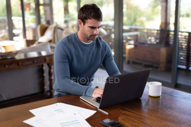 Kaukasier sitzt am Tisch und arbeitet mit Laptop. Arbeiten zu Hause in moderner Wohnung. — Stockfoto