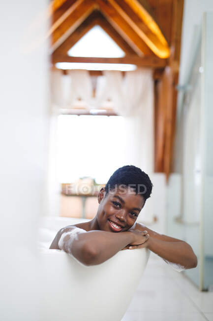 Portrait de femme afro-américaine souriante dans la salle de bain, relaxante dans la baignoire. mode de vie domestique, profiter de loisirs d'auto-soins à la maison. — Photo de stock