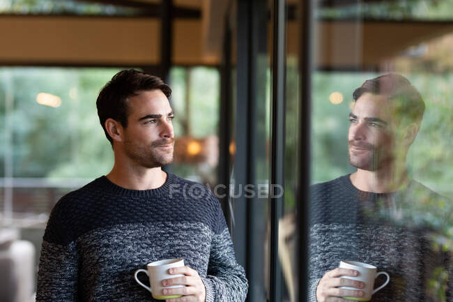 Caucasian man in living room looking through window holding mug and smiling. spending time off at home in modern apartment. — Stock Photo