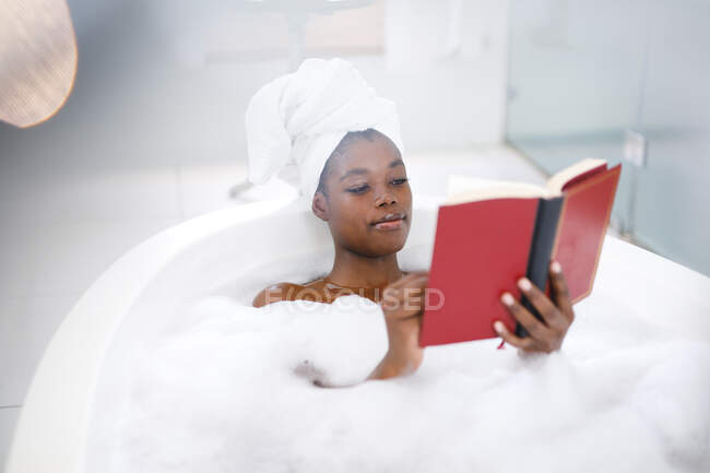 Feliz mulher afro-americana no banheiro relaxante em livro de leitura de banho. estilo de vida doméstico, desfrutando de tempo de lazer auto-cuidado em casa. — Fotografia de Stock
