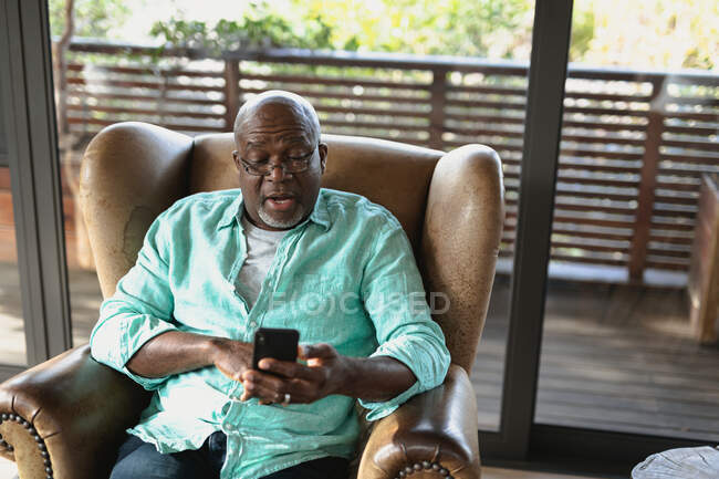 Hombre afroamericano mayor sentado en el sillón y utilizando el teléfono inteligente en la sala de estar moderna. estilo de vida de jubilación, pasar tiempo solo en casa. - foto de stock