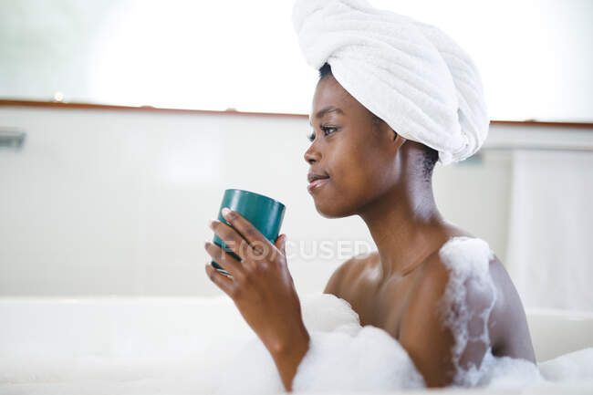 Femme afro-américaine souriante dans la salle de bain, se détendre dans la baignoire et boire du café. mode de vie domestique, profiter de loisirs d'auto-soins à la maison. — Photo de stock