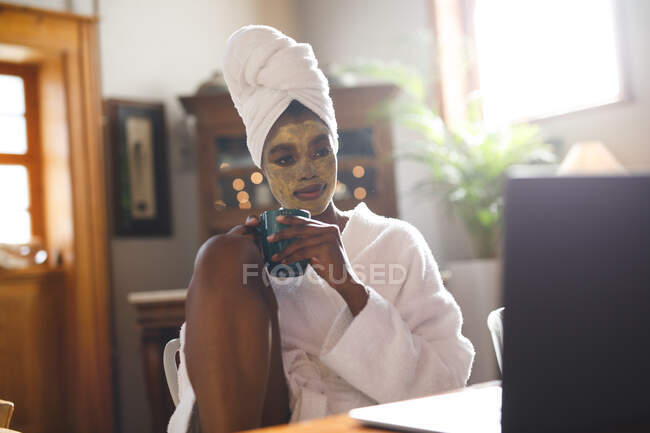 Mujer afroamericana sonriente con mascarilla de belleza en la sala de estar usando computadora portátil y tomando café. estilo de vida doméstico, disfrutando del tiempo libre de autocuidado en casa. - foto de stock