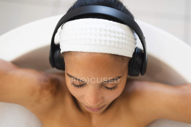 Mulher de raça mista no banheiro relaxando no banho usando fones de ouvido, com os olhos fechados. estilo de vida doméstico, desfrutando de tempo de lazer auto-cuidado em casa. — Fotografia de Stock