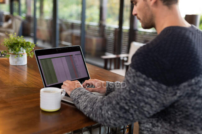 Homem caucasiano sentado à mesa na cozinha trabalhando remotamente usando laptop. em casa no apartamento moderno. — Fotografia de Stock