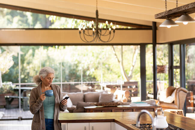 Senior kaukasische Frau in der Küche mit Smartphone und Kaffee trinken. Lebensstil im Ruhestand, Zeit allein zu Hause verbringen. — Stockfoto