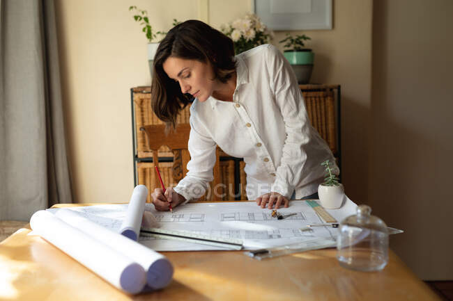 Arquiteta branca na sala de estar, sentada à mesa a trabalhar, a desenhar planos. estilo de vida doméstico, trabalho remoto de casa. — Fotografia de Stock