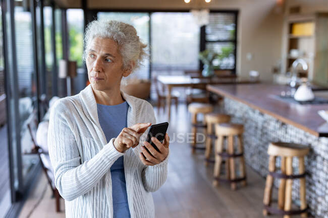 Старшая белая женщина на кухне стоит и пользуется смартфоном. пенсионного образа жизни, проводить время в одиночестве на дому. — стоковое фото