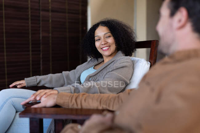 Feliz pareja diversa en la sala de estar sonriendo y tomados de la mano. pasar tiempo libre en casa en apartamento moderno. - foto de stock