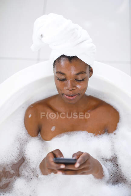 Femme afro-américaine souriante dans la salle de bain, se détendre dans la salle de bain à l'aide d'un smartphone. mode de vie domestique, profiter de loisirs d'auto-soins à la maison. — Photo de stock