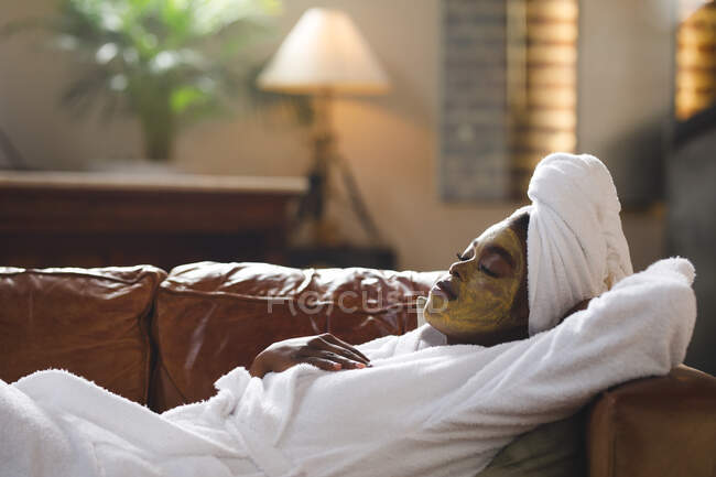Donna afroamericana sorridente con maschera viso di bellezza in soggiorno sdraiata sul divano con gli occhi chiusi. stile di vita domestico, godendo di auto cura del tempo libero a casa. — Foto stock