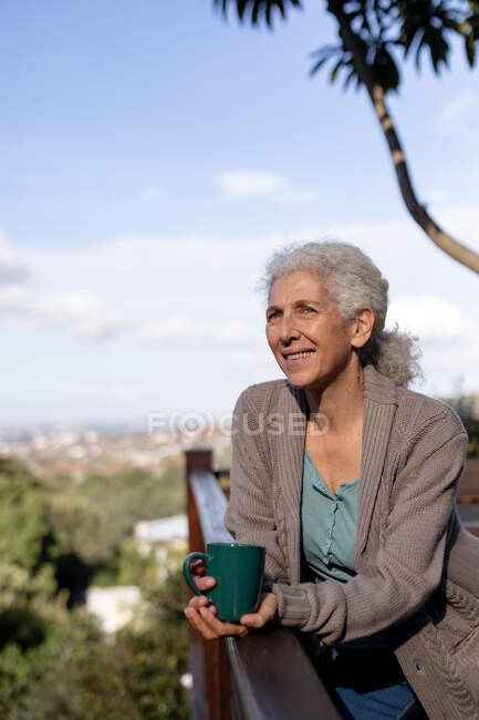 Rilassante donna caucasica anziana sul balcone in piedi e bere caffè. stile di vita di pensione, trascorrere del tempo da solo a casa. — Foto stock