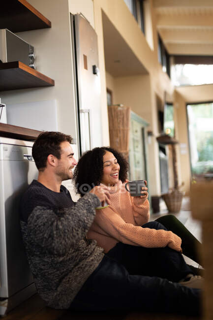 Happy diverse couple in kitchen sitting on floor and drinking coffee. spending time off at home in modern apartment. — Stock Photo