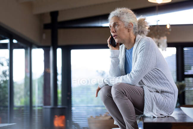 Eine ältere kaukasische Frau in der Küche sitzt auf dem Tisch und benutzt ihr Smartphone. Lebensstil im Ruhestand, Zeit allein zu Hause verbringen. — Stockfoto