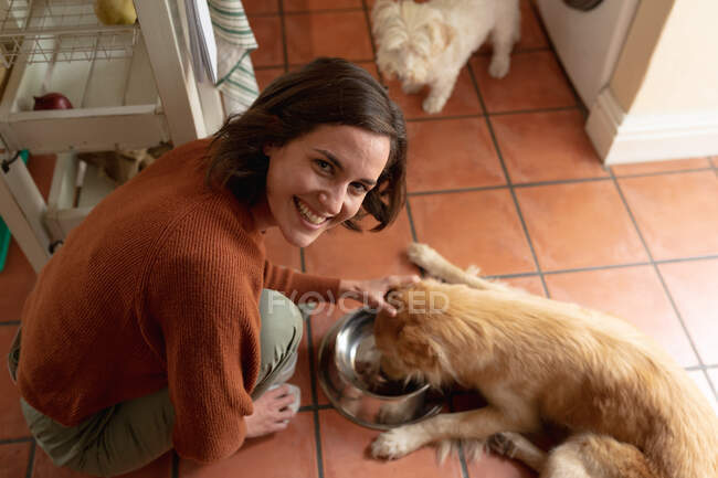 Portrait of smiling caucasian woman in kitchen feeding pet dogs. domestic lifestyle, enjoying leisure time at home. — Stock Photo