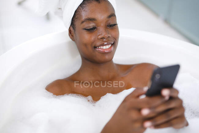 Sorrindo mulher afro-americana no banheiro, relaxando no banho usando smartphone. estilo de vida doméstico, desfrutando de tempo de lazer auto-cuidado em casa. — Fotografia de Stock