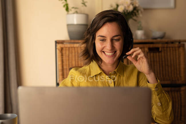 Smiling caucasian woman in living room, sitting at table working, using laptop wearing headset. domestic lifestyle, remote working from home. — Stock Photo