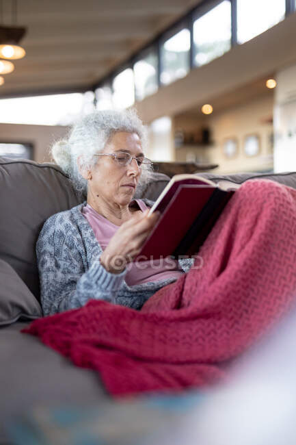 Senior caucasian woman sitting on the couch and reading book in the modern living room. retirement lifestyle, spending time alone at home. — Stock Photo