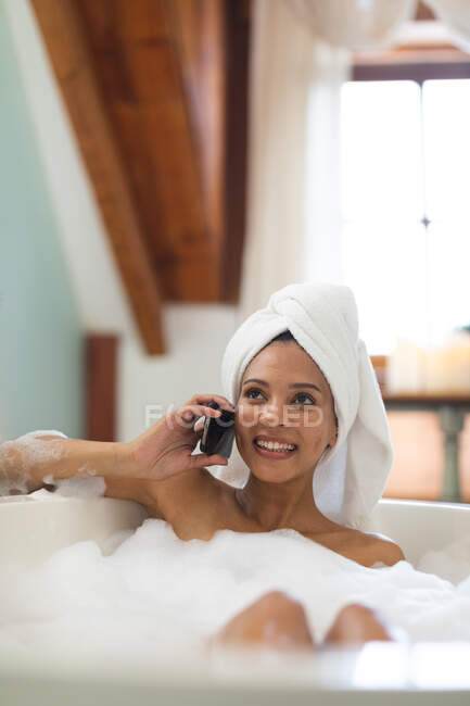 Femme métissée souriante dans la salle de bain ayant une baignoire et parlant sur smartphone. mode de vie domestique, profiter de loisirs d'auto-soins à la maison. — Photo de stock