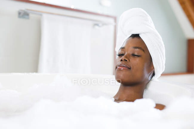 Улыбающаяся африканская американка в ванной комнате расслабляется в ванной с закрытыми глазами. домашний образ жизни, наслаждаясь отдыхом на дому. — стоковое фото