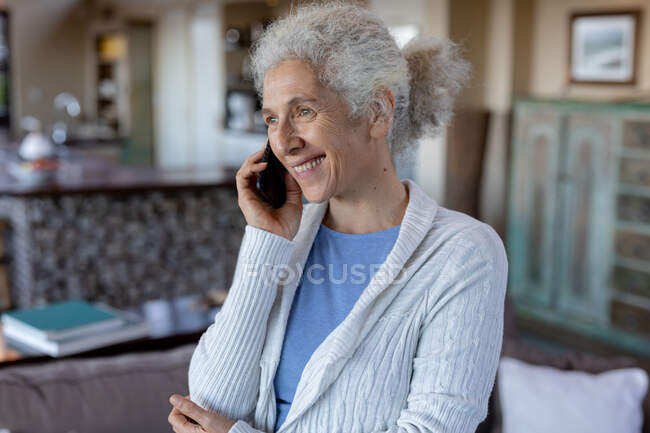 Donna caucasica anziana in soggiorno in piedi e utilizzando il telefono cellulare. stile di vita di pensione, trascorrere del tempo da solo a casa. — Foto stock