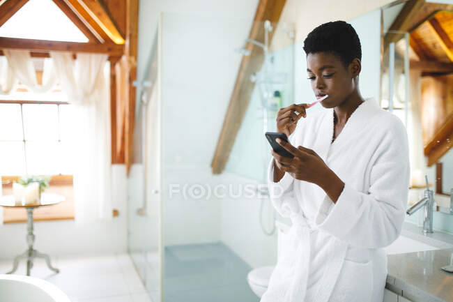 African american woman in bathroom brushing teeth and using smartphone. domestic lifestyle, enjoying self care leisure time at home. — Stock Photo