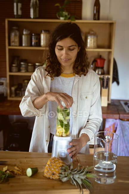 Смешанная расовая женщина готовит здоровый напиток на кухне. здоровый образ жизни, наслаждаясь отдыхом дома. — стоковое фото