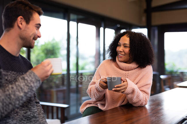 Happy diverse couple in kitchen sitting at table drinking coffee and talking. spending time off at home in modern apartment. — Stock Photo
