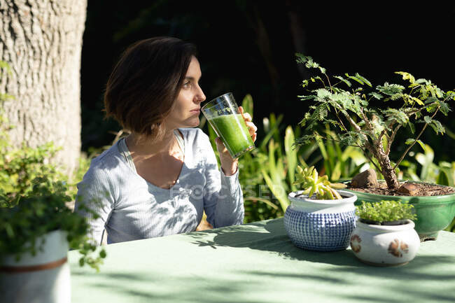 Белая женщина в саду, сидит за столом и пьет здоровый напиток. домашний образ жизни, наслаждаясь отдыхом дома. — стоковое фото
