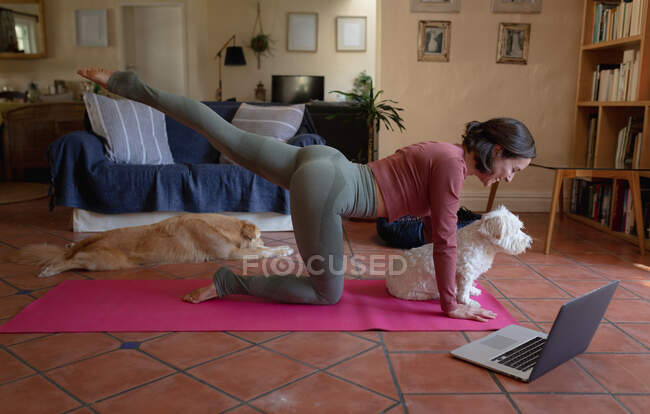 Lächelnde Kaukasierin im Wohnzimmer mit ihren Vierbeinern, Yoga praktizierend, mit Laptop. häuslicher Lebensstil, Freizeit zu Hause genießen. — Stockfoto