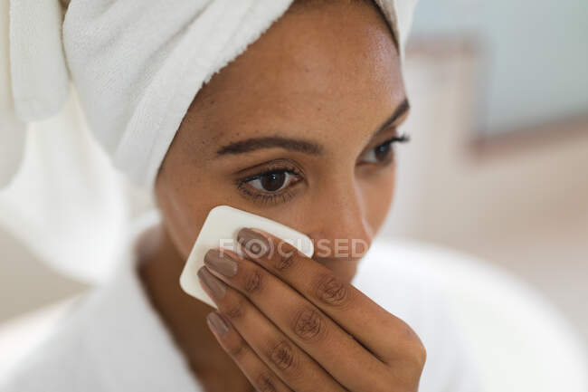 Mixed race woman in bathroom cleansing her face with cotton pad for skin care. domestic lifestyle, enjoying self care leisure time at home. — Stock Photo