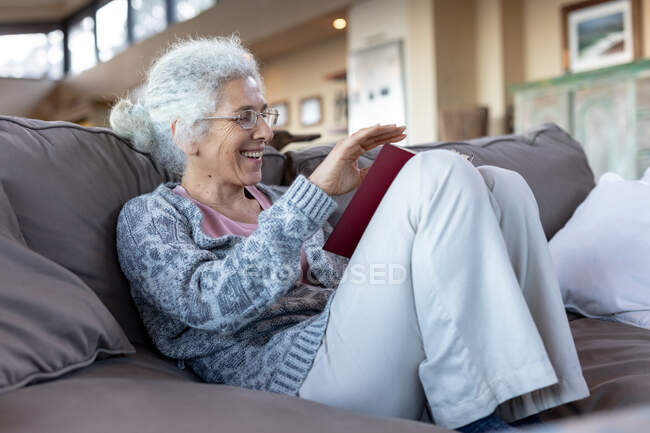 Felice donna caucasica anziana seduta sul divano e leggere libro nel soggiorno moderno. stile di vita di pensione, trascorrere del tempo da solo a casa. — Foto stock