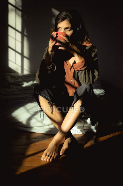 Mujer de raza mixta sentada y tomando café en el dormitorio soleado. estilo de vida saludable, disfrutando del tiempo libre en casa. - foto de stock