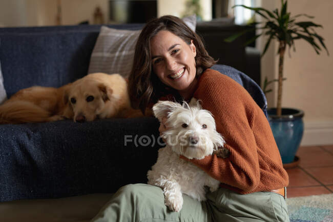 Retrato de una mujer caucásica sonriente en la sala de estar sentada en el suelo abrazando a su perro mascota. estilo de vida doméstico, disfrutando del tiempo libre en casa. - foto de stock