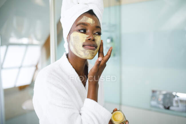 Sorrindo mulher afro-americana no banheiro aplicando máscara facial beleza. estilo de vida doméstico, desfrutando de tempo de lazer auto-cuidado em casa. — Fotografia de Stock