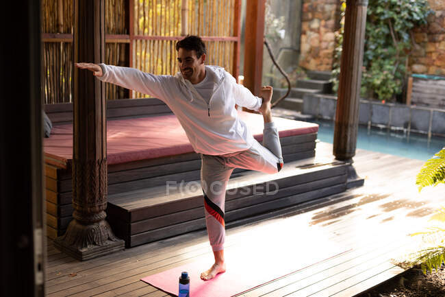 Hombre caucásico usando ropa deportiva y practicando yoga de pie en la esterilla de yoga. pasar tiempo libre en casa. - foto de stock